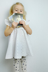 close-up of small child, elegant blonde girl 2 years old in white dress holds a stack of old...