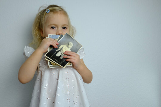 close-up of small child, elegant blonde girl 2 years old in white dress holds a stack of old photographs of 1950-1960, the concept of family tree, childhood, family connection of generations