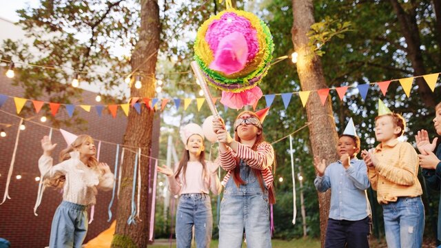 Portrait of small girl smash pinata with friends and presents outdoors in summer. Happy birthday concept