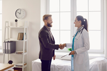Fototapeta premium Happy grateful male patient shakes hands with his female doctor at meeting in hospital office. Smiling man and young female doctor or nurse have pleasant conversation standing in examination room.
