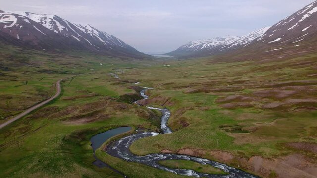 Aerial shot flying through green valley over river with snow covered mountains surrounding. Shot in Iceland. Epic landscape scenery.