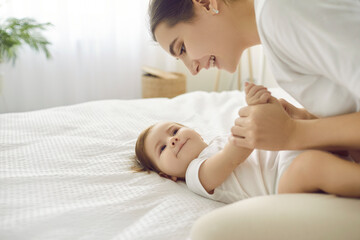 Happy mother playing with her baby. Charming infant looking at mom with cute, funny face expression. Beautiful young mommy and her little child cuddling on clean white bed in nursery room or bedroom