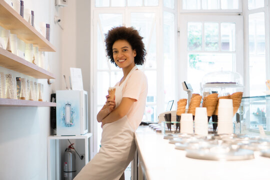 A beautiful mixed-race woman is smiling behind the stand of an ice cream parlor.