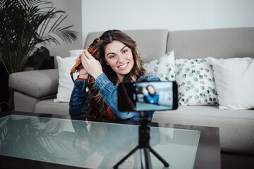 Smiling hairstylist with straightener curling hair and filming through mobile phone at home