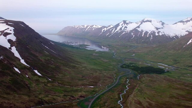 High aerial shot with view of Icelandic fjord. Lush green below and coastal town in the distance. Snow covered mountains, epic scenery. 