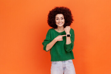 Good looking positive woman with Afro hairstyle wearing green casual sweater pointing at wristwatch...