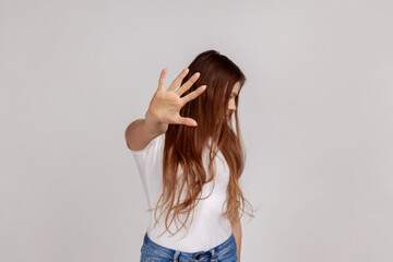Fototapeta premium Portrait of dark haired woman showing stop sign with outstretched palm, does not want to see, prohibition ban gesture, wearing white T-shirt. Indoor studio shot isolated on gray background.