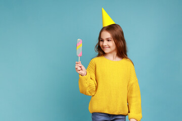 Portrait of cute little girl in party cone standing with delicious ice cream, expressing happiness, wearing yellow casual style sweater. Indoor studio shot isolated on blue background.