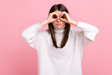 Portrait of attractive brunette female standing raised arms and looking with binoculars gesture, wearing white casual style sweater. Indoor studio shot isolated on pink background.