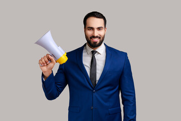 Fototapeta premium Bearded young adult man with satisfied expression, holding megaphone in hands, looking at camera with toothy smile, wearing official style suit. Indoor studio shot isolated on gray background.
