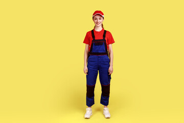 Fototapeta na wymiar Full length portrait of worker woman standing and looking at camera with positive facial expression, wearing overalls and red cap. Indoor studio shot isolated on yellow background.