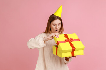 Portrait of excited amazed blond woman in party cone unpacking present box, having pleasant surprise, being shocked, wearing white sweater. Indoor studio shot isolated on pink background.