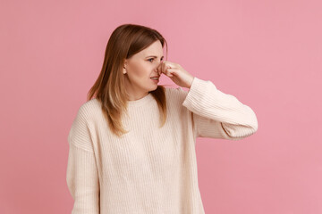 Portrait of blond woman grimacing with disgust, holding breath, pinching nose with fingers to avoid bad smell, awful odor, wearing white sweater. Indoor studio shot isolated on pink background.