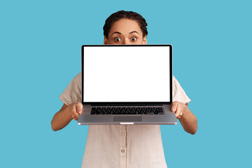 Portrait of unknown woman with dreadlocks covering half of face with laptop with empty display for advertisement or promotional text, wearing shirt. Indoor studio shot isolated on blue background.