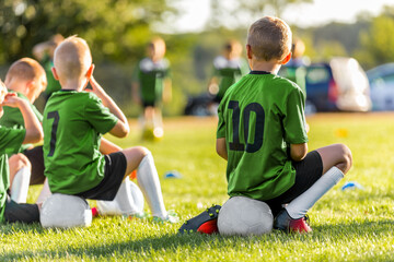 Obraz premium Group of Young Boys Kicking Sports Soccer Game on Grass Pitch. Boys Sitting on Soccer Balls Waiting to Play the Game. Friends in Sports Team. School Boys on Sideline. Kids in Green-Sports Shirts
