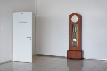 A large modern grandfather clock with weights and pendulum in a white empty room.