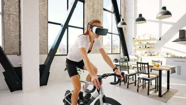 Young female in VR glasses is pedaling on indoor exercise bike trainer. Studio flat, kitchen. Daily training at home. Slow motion