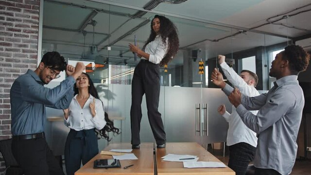 Multiracial employees have fun rejoice together celebrating success confident young girl dancing on desk in office woman boss hold folder in hand enjoy victory colleagues in boardroom corporate party