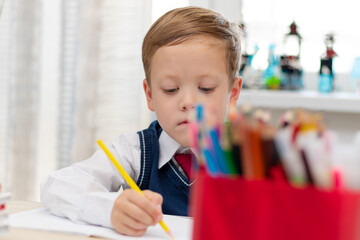 A cute boy first grader in a school uniform does homework while sitting at a desk with a pencil in...