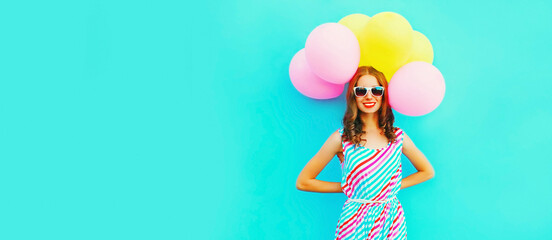 Portrait of beautiful happy smiling young woman with bunch of balloons wearing a colorful dress on blue background