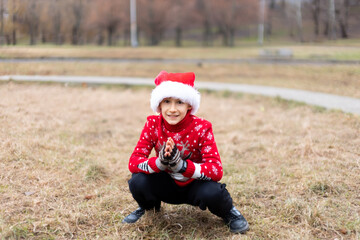 a boy in a warm Christmas sweater with a New Year's deer and a Santa hat crouched down and looks into the camera