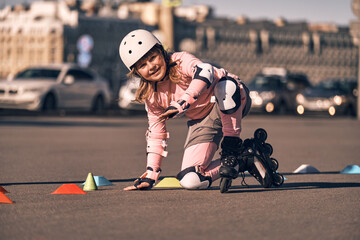 Happy child learns to roller skate on the streets of the city next to the cars. Girl dressed in...