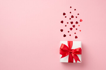 Top view photo of st valentine's day decorations white giftbox with red ribbon bow and red heart shaped confetti on isolated pastel pink background with copyspace