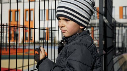 Abused and stressed little boy holding metal fence and looking on children palyground