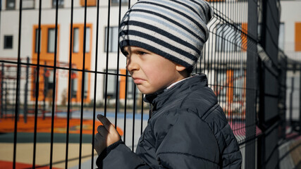 Upset lonely boy lenaing and holding metal net fence at school playground