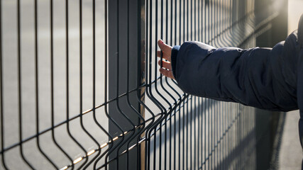 Closeup of little boy walking by the metal fence and touching it with hand. Concept of poverty,...