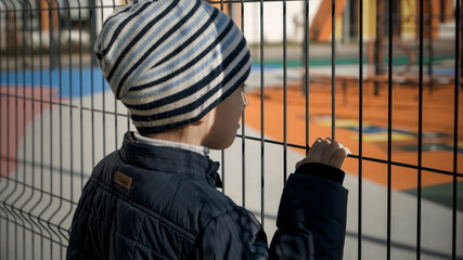 Upset stressed little boy holding and looking through high metal fence on expensive school and playground. Concept of poverty, immigration, bullying and kids stress