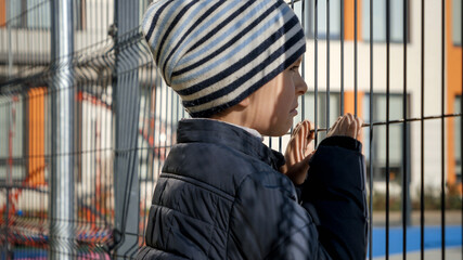 Upset and stressed boy looking through school metal fence after was bullied. Concept of poverty, immigration, bullying and kids stress