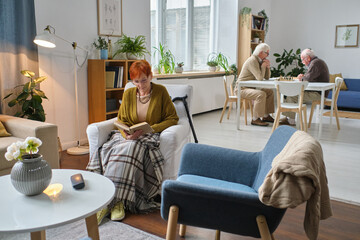 Elderly woman sitting in armchair in the room and reading a book with men playing chess in the...