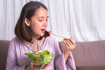 Happy Pregnant woman holding glass bowl with fresh salad. Eating healthy vegetable organic food for baby. Care about unborn child. sitting on sofa in living room. Copy space for text.