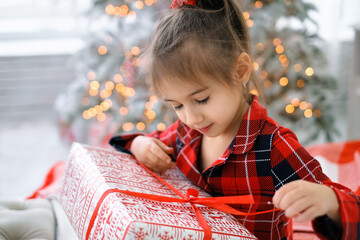 Cute girl unties a red bow on a Christmas gift while sitting on a bed by a Christmas tree with...