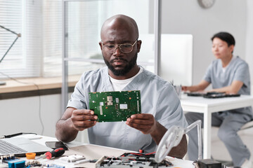 Serious African man in workwear looking at motherboard in hands while repairing it in workshop