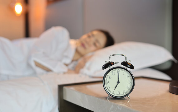 Alarm clock with blurred image of oversleeping woman wearing white sleepwear lying or sleeping on bed in bedroom with happiness and smile. Lifestyle concept