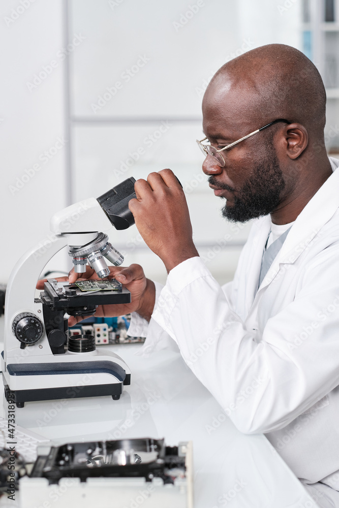 Poster Young serious African researcher looking in microscope while studying new microchip by desk in scientific laboratory