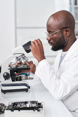 Young serious African researcher looking in microscope while studying new microchip by desk in scientific laboratory