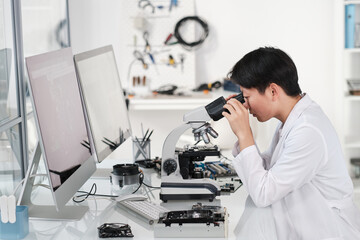 Young Asian female researcher looking in microscope while making scientific investigation of hardware
