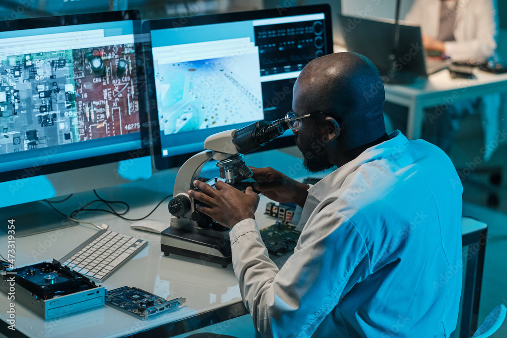 Poster Contemporary young African scientist with microscope working in front of computer monitors