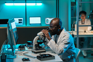 Young African laboratory worker looking in microscope during scientific investigation