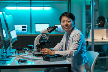 Young successful chinese female researcher with microscope looking at camera by workplace in laboratory