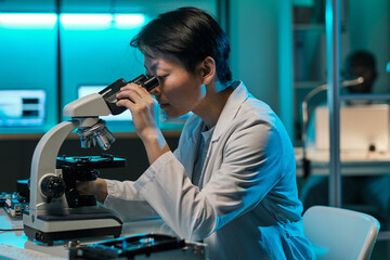 Young serious female laboratory worker in whitecoat looking at microchip in microscope while...