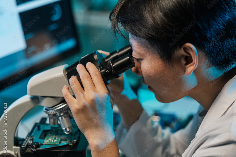 Poster Young Asian technician looking in microscope while studying microchip characteristics in laboratory