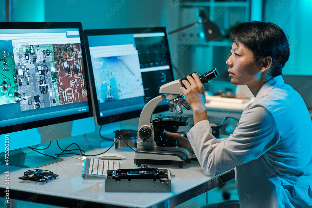 Poster young serious female scientist with microscope looking at computer screen while sitting by workplace