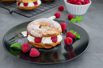 Choux pastries. Choux rings with cream of cream cheese or cottage cheese and fresh raspberries, dusted with powdered sugar on a black plate on a gray concrete background.