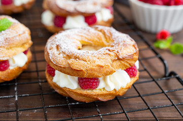 Choux pastries. Choux rings with cream of cream cheese or cottage cheese and fresh raspberries, dusted with powdered sugar on a dark wooden background.
