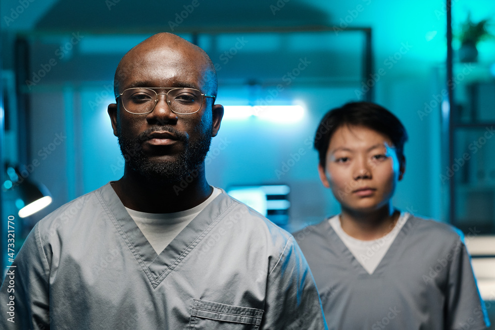 Poster two young intercultural researchers in uniform looking at camera while standing in laboratory