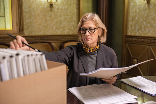 Mature blond lawyer in formalwear revising juridical documents in folders while sitting by table in boardroom